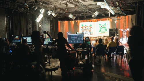 Photo of the Conjuring StoryArcs Demo in the Futures Studio, by Alex Grigoras. An audience in a dimly lit studio faces an illuminated virtual production wall displaying an open storybook, whilst the tech team operates several computers behind them.