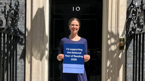 Jessie Ricketts, woman with brown hair smiling, standing in front of black door to Downing Street holding a piece of card reading "I'm supporting the National Year of Reading 2026